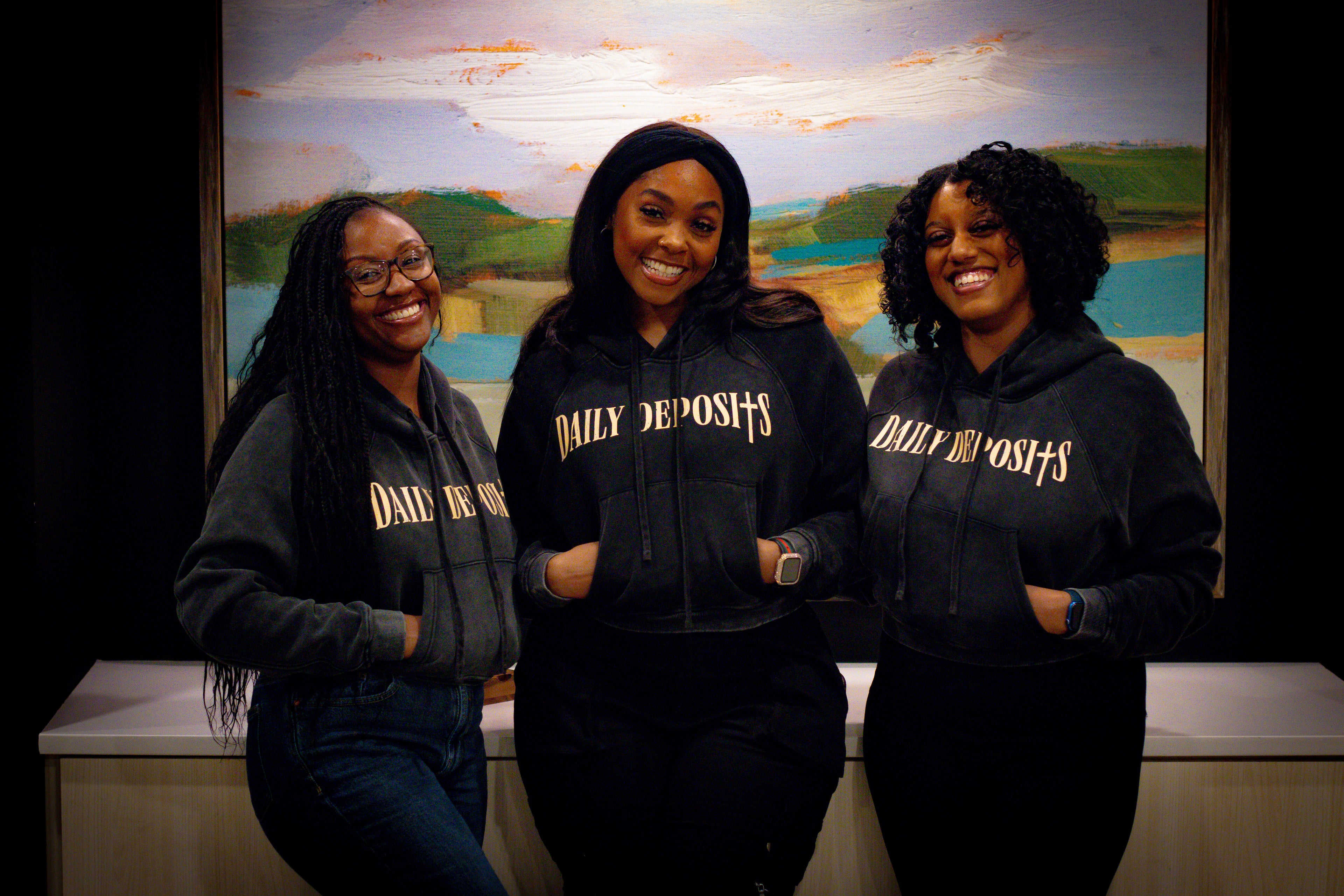 Three women wearing black hoodies with 'Daily Deposits' text in front of a scenic painting.