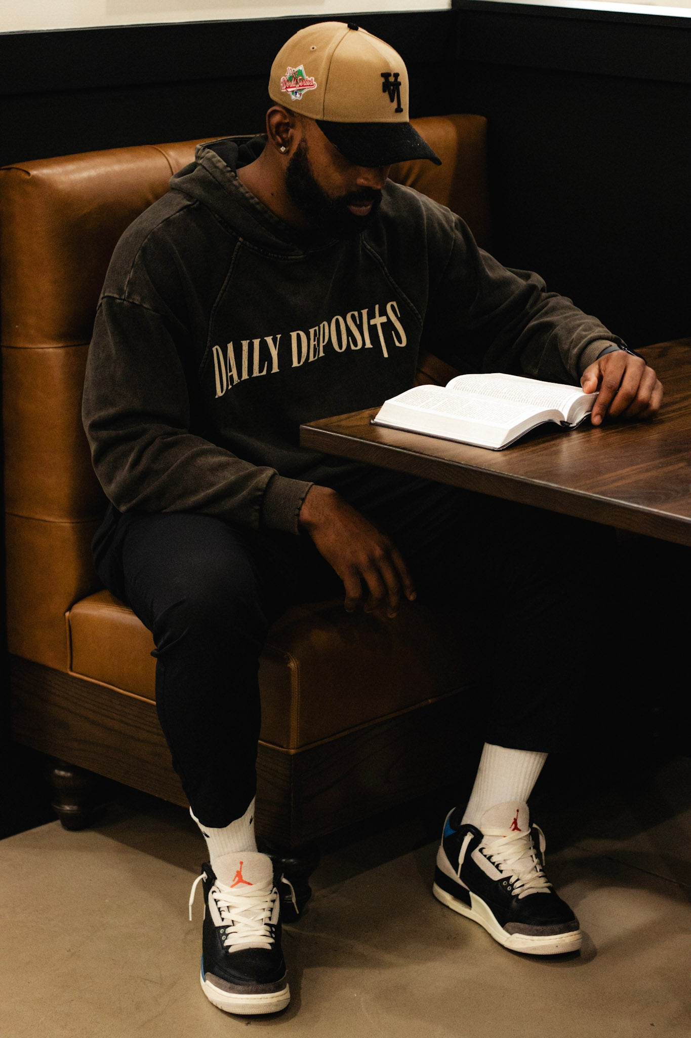 Man sitting at a table with an open book, wearing a 'Daily Deposits' sweatshirt and a cap.